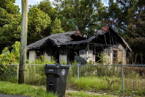 First floods, now fires: How neglect and fraud hobbled Prichard A burned-out house, its roof caved in and its top turned to blacked cinders. Plants and grass grown around it. A black trash sits in the foreground, outside a fence.