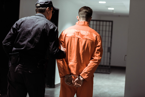 Constitutional amendment to tighten money bail system passes Assembly rear view of prison officer leading prisoner in handcuffs in corridor in jail. Inmate wearing orange