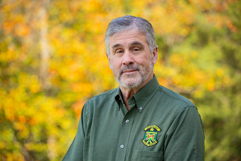 Wild divide: A debate over wildlife management in Vermont runs deep An older man with gray hair and a beard, wearing a green shirt with a badge, smiling gently against a blurred autumnal background.